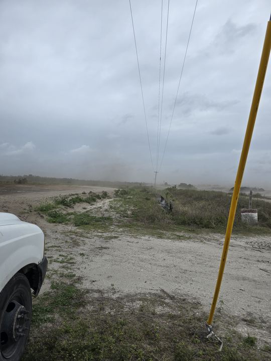 Truck parked off the road beside an open sandy field, with lines overhead and a yellow pole guard