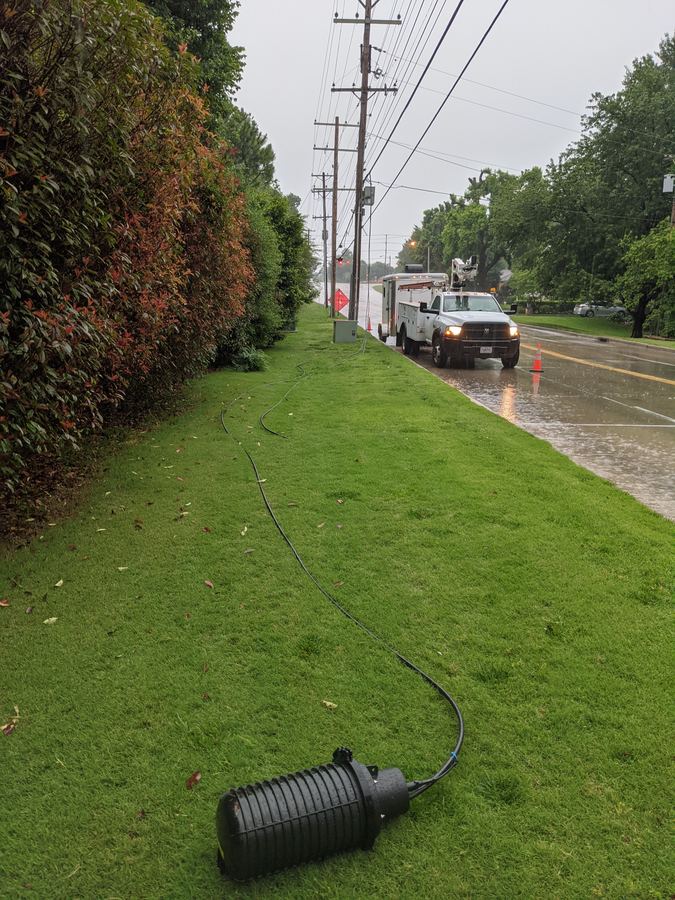 Bucket and splice truck set up on a wet road in the rain with cones and an orange warning sign, with a closure and cable laid out on the grass
