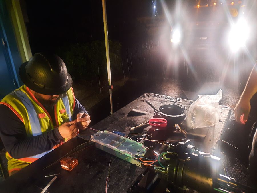 Splicer in a hard hat and hi-vis vest working a splice tray on a tailgate at night in the rain, lit by truck headlights