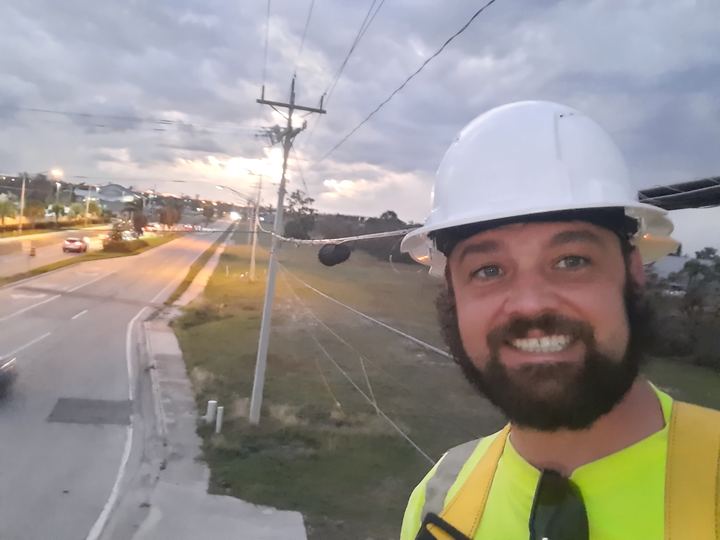 Crew member in a white hard hat and hi-vis vest beside a roadside pole with sagging lines under stormy evening skies