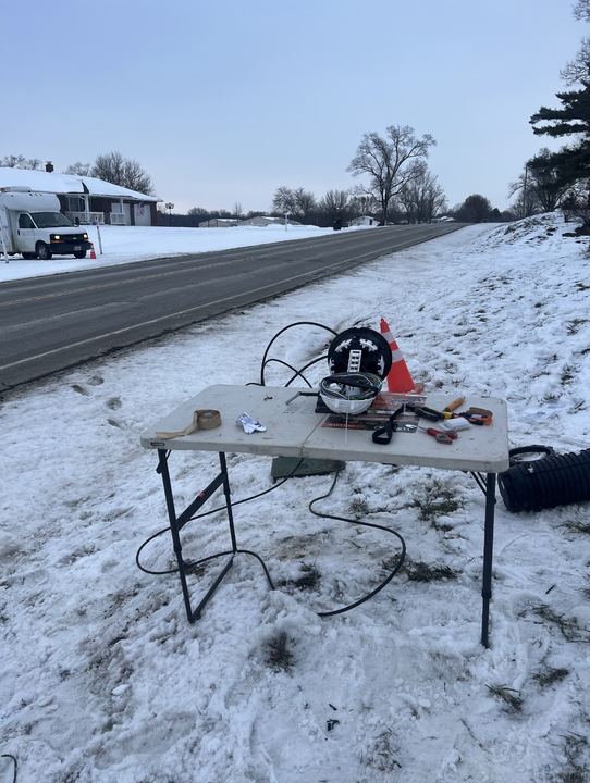 Roadside splicing table in the snow with a cone, a closure and tools, and a splice truck in the background